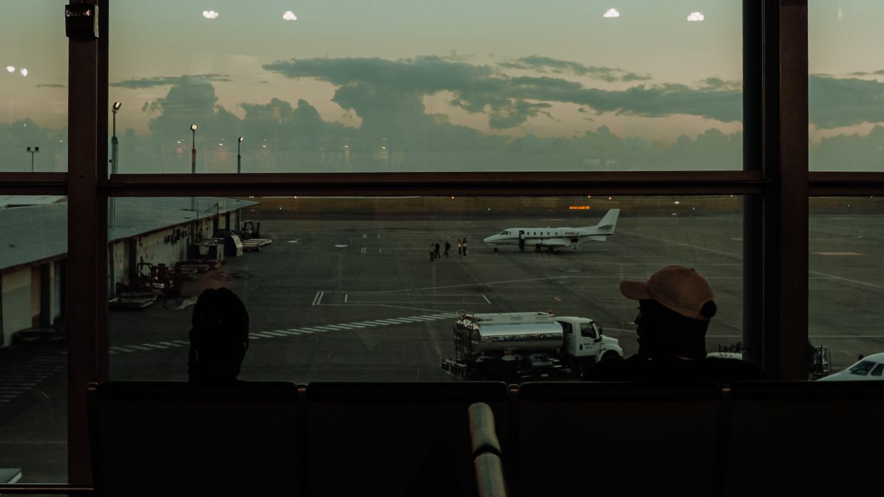 Travellers silhouetted at a terminal window at dusk, an aircraft on the apron beyond